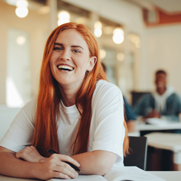 a girl with red hair sitting at a desk, holding a pen and smiling